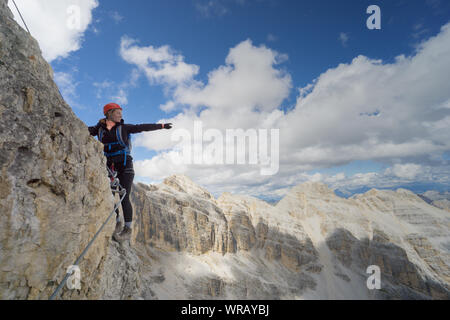 Horizontal view of attractive brunette female climber on a steep and exposed Via Ferrata in the Dolomites pointing to the distance Stock Photo