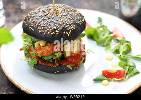 Burger in a black bun with chopped meat, pickled cucumber and ketchup served on a plate. Stock Photo
