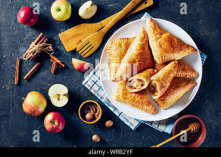 apple turnover on a white plate on a dark concrete table with honey in a bowl and ingredients, horizontal view from above, flatlay, free space Stock Photo
