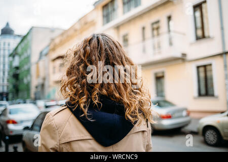 Woman with curly hair in a beige coat with a hood walks around the city, rear view Stock Photo