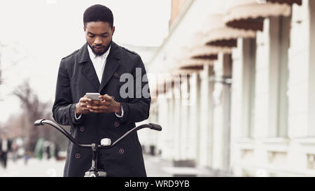 Afro businessman texting on phone, standing with bike outdoors Stock Photo