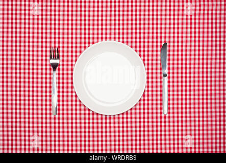 Place setting, round white plate, knife and fork, from above on a red gingham tablecloth Stock Photo