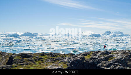 Travel wanderlust adventure in Arctic landscape nature with icebergs - tourist person looking at view of Greenland icefjord - aerial photo. Man by ice and iceberg, Ilulissat Icefjord. Stock Photo