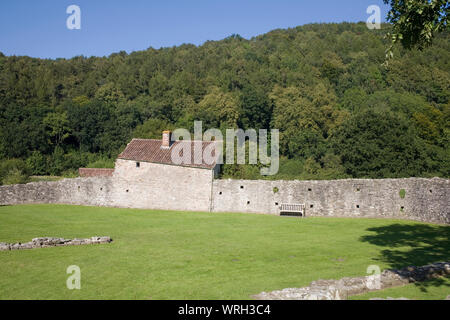 Walls surrounding Tintern abbey with stone walled cottage, grass area and wooden bench with forested hillside beyond Stock Photo