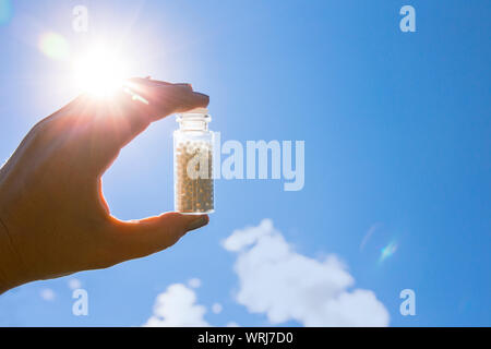 Selective focus on person hand holding glass jar full of small white round homeopathy pills against blue sky background. Stock Photo