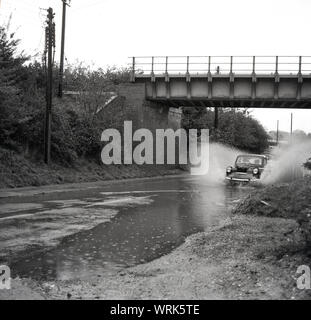 1967, historical, flooded road, car of the era going under a bridge through a large pool of water on a road where the drains have overflowed. Risborough, England, UK. Stock Photo