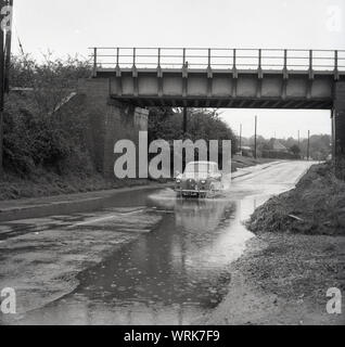 1967, historical, an Austin morris car going under a bridge through a large pool of water on a flooded road, England, UK. Stock Photo