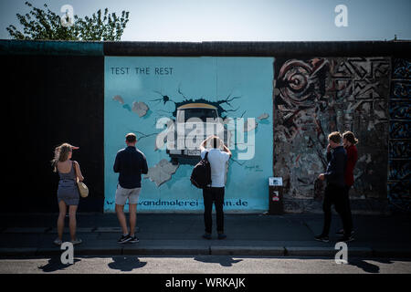 22 August 2019, Berlin: Visitors and tourists stand in front of the Trabi picture of the artist Birgit Kinder at the East Side Gallery. Almost 30 years ago, artists immortalized themselves here and reinterpreted the longest remaining section of the Berlin Wall with their works of art. (for 'Wall Artist Avignon: East Side Gallery awarded anew each year') Photo: Arne Immanuel Bänsch/dpa Stock Photo