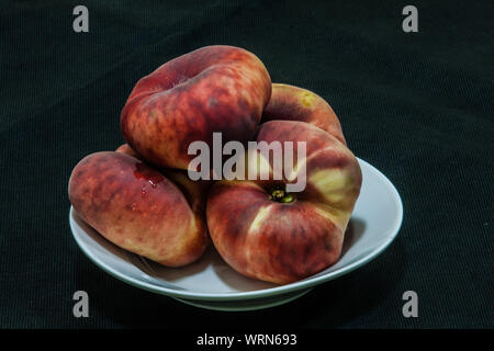 Flat peaches (Prunus persica var. platycarpa) in a deep plate on the dark background Stock Photo