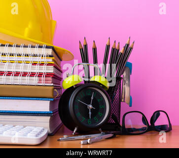 Close up educational materials on wooden desk, construction helmet, stack of book, calculator, pencils, clock, dividers, stapler and glasses isolated Stock Photo