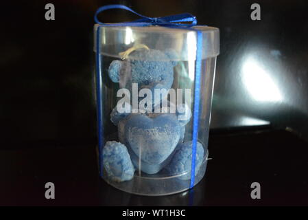 Blue teddy bear made of natural wax is made in the shape of a candle and packed in a plastic box with blue bow on black glossy background. Stock Photo