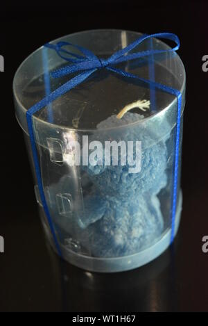 Blue teddy bear made of natural wax is made in the shape of a candle and packed in a plastic box with blue bow on black glossy background. Stock Photo