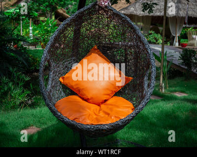 Modern black rattan lounger hanging egg chair with orange pillow in the garden. Stock Photo