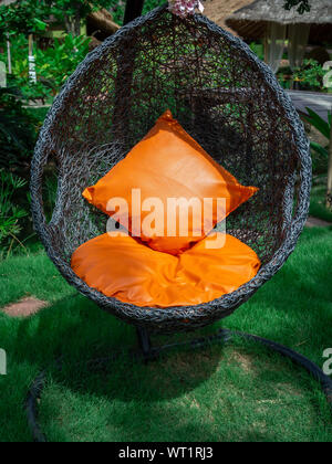 Modern black rattan lounger hanging egg chair with orange pillow in the garden vertical style. Stock Photo