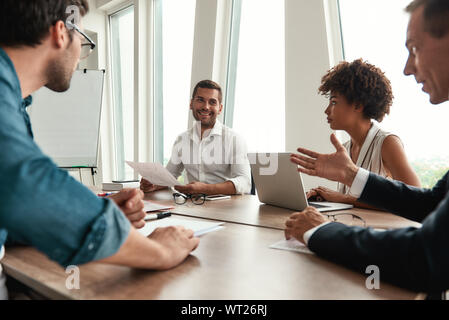 Analyzing results. Young man in formal wear holding financial reports and discussing it with team while sitting at the office table. Business people. Brainstorming Stock Photo