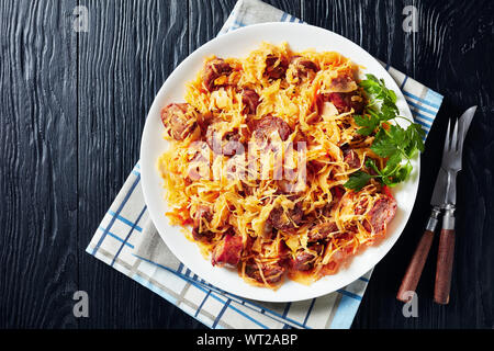 delicious Cabbage stew with sliced fried sausages served on a white plate on a black wooden table, view from above, flat lay, close-up Stock Photo