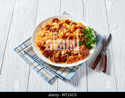 homemade Cabbage stew with sliced fried sausages served on a white plate on a wooden table with fork and knife, view from above Stock Photo