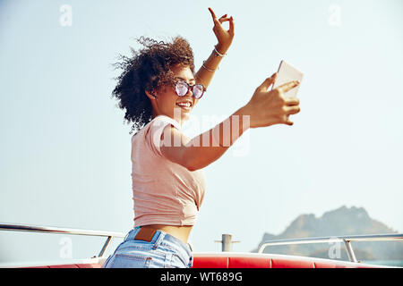 Carefree young woman wearing sunglasses standing on a boat against a blue sky taking selfies during her summer vacation Stock Photo