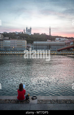 LYON, FRANCE - JULY 17, 2019: Young woman reading a book & relaxing on the riverbank of Saone river in Lyon, at dusk, with Colline and Basilique Fourv Stock Photo