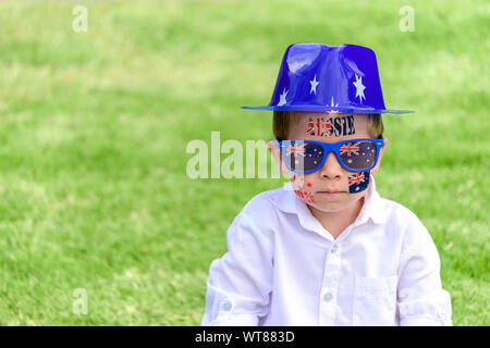 Serious Australian boy with sunglasses and hat sitting on lawn during Australia Day celebration Stock Photo