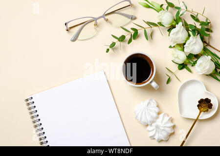Flat lay feminine accessories, glasses, blank paper notepad, coffee cup, marshmallow and white roses flowers on pastel colors beige background. Top vi Stock Photo