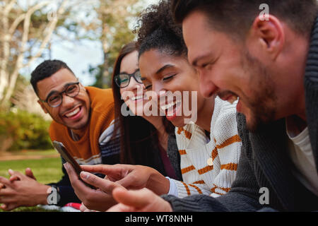 group of multi ethnic friends in college lying together in the park enjoying watching text or video on mobile phone - smiling group of students Stock Photo