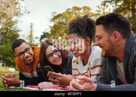 Group of happy diverse friends looking at african american woman using her mobile phone in the park on a summers day - very happy young group of Stock Photo