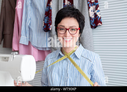 Smiling dressmaker in eyeglasses working at tailor shop with electrical industrial sewing machine Stock Photo