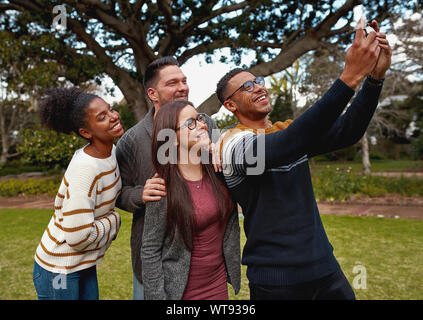 Group of multiracial young friends standing together in a green park taking a selfie on mobile phone smiling - a summer picnic  Stock Photo