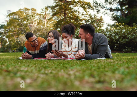 Smiling african american young woman student showing something interesting to her friends lying together on blanket over the green grass in park Stock Photo