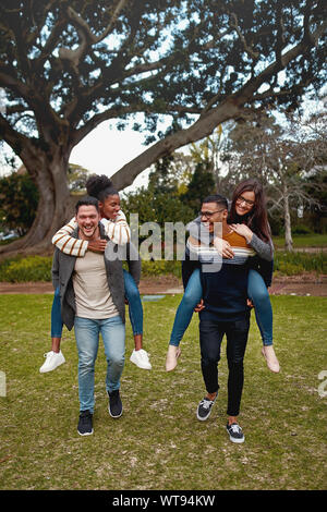 Happy young men carrying piggyback ride to their girlfriends in a park smiling - having fun outdoors Stock Photo