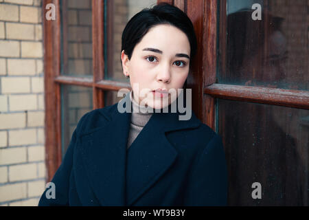 Close up portrait of a fashionable brunette woman with short hair and wearing a blue coat on the background of a door frame. Adorable millennial girl Stock Photo