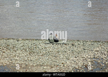 Young couple sitting at the edge of the river Thames on a pipe at low tide on a day in summer. Stock Photo