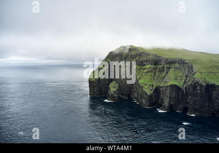 Rough rocky cliff covered with green grass located near rippling sea on overcast day on Faroe Islands Stock Photo