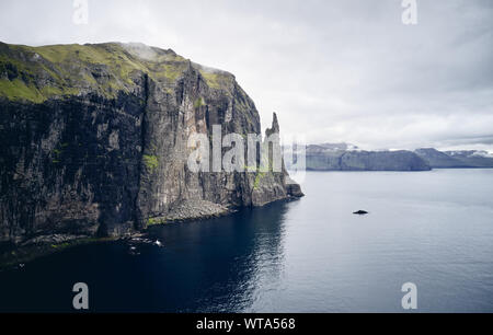 Majestic rocky cliff with weathered surface located near tranquil sea against gray overcast sky on Faroe Islands Stock Photo