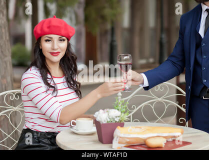 Elegant man in a suit gives a pretty brunette woman in a red cap and striped t-shirt glass of red wine in outdoor cafe. Stock Photo