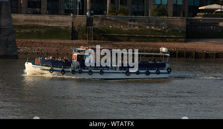 Small tourist cruise boat, the Sarah Kathleen,  going upstream into  the evening sun on the river Thames at low tide near the gravel of the river bank Stock Photo