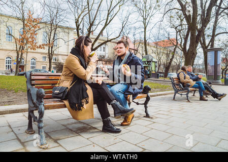 young adult couple eating fast food on bench of city park Stock Photo
