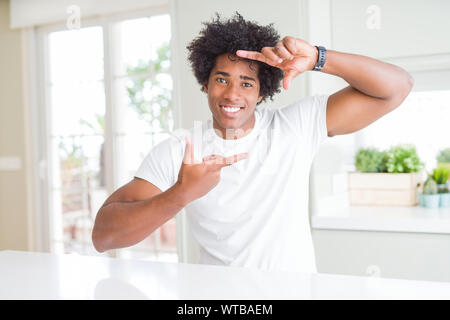 Young african american man wearing casual white t-shirt sitting at home smiling making frame with hands and fingers with happy face. Creativity and ph Stock Photo