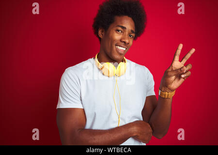 Young african american man listening to music using headphones over isolated red background smiling with happy face winking at the camera doing victor Stock Photo