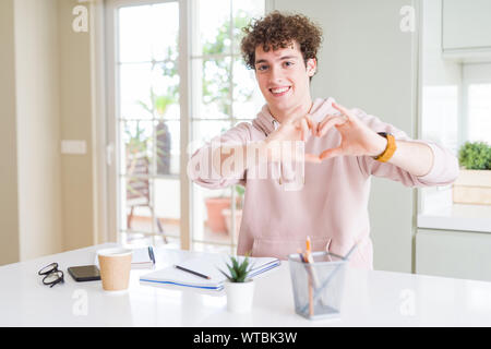 Young student man writing on notebook and studying smiling in love showing heart symbol and shape with hands. Romantic concept. Stock Photo