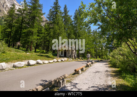 Scenic view of an Alpine mountain road among forests with a small church and a cyclist in summer, Val Ferret, Courmayeur, Aosta Valley, Alps, Italy Stock Photo