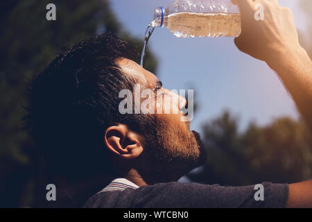 Young man splashing and pouring fresh water from a bottle on his head to refresh against a blue sky background in a summer heat Stock Photo