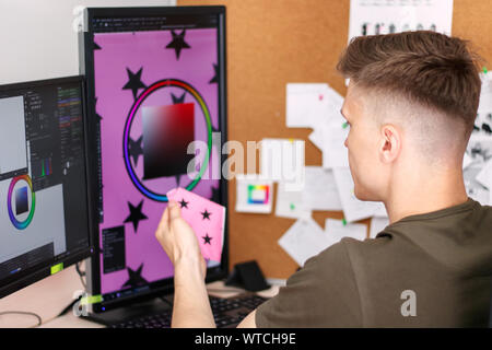 Side view of a young male retoucher checks the color of the product in the photo with a sample of fabric Stock Photo