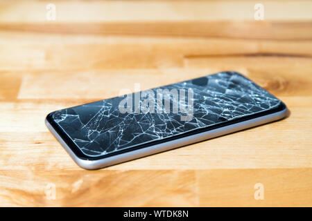 Concept of smart phone with broken screen. Top view on wooden desk background. Cracked, shattered lcd touch screen on modern cellphone. Gadget needs r Stock Photo
