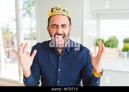 Handsome hispanic man wearing golden crown over head as the king celebrating mad and crazy for success with arms raised and closed eyes screaming exci Stock Photo