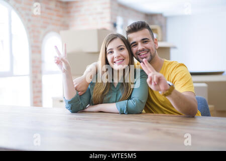 Young couple sitting on the table movinto to new home with carboard boxes behind them smiling with happy face winking at the camera doing victory sign Stock Photo