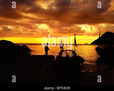 Silhouette Man On Pier By Boat In Sea Against Dramatic Cloudy Sky During Sunset