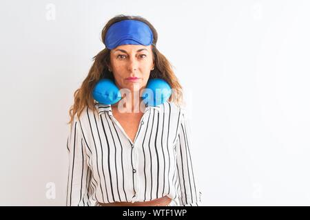 Middle age tourist woman wearing neckpillow and sleep mask over isolated white background with serious expression on face. Simple and natural looking Stock Photo