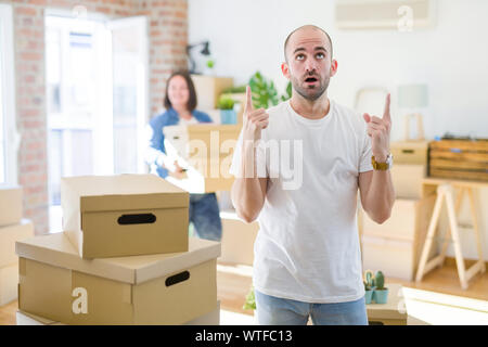 Young couple arround cardboard boxes moving to a new house, bald man standing at home amazed and surprised looking up and pointing with fingers and ra Stock Photo